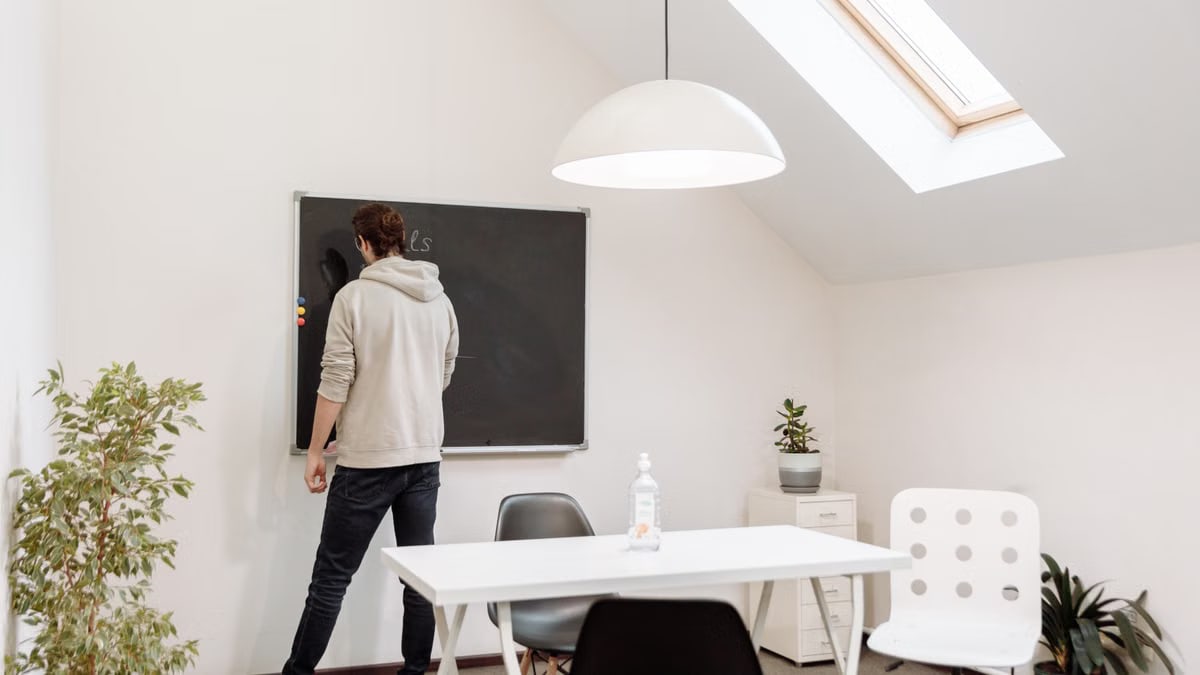 A person in a hoodie writes on a blackboard in a minimalist office with a skylight.
